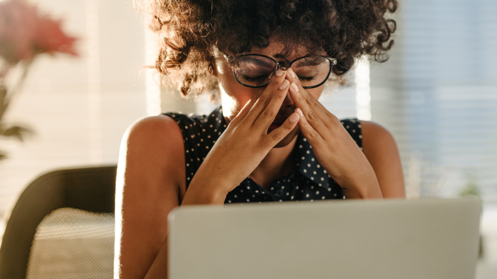 black woman with curly hair rubbing eyes while looking at computer